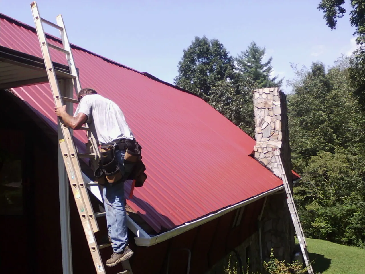 Roofer on a ladder beside a red metal roof and stone chimney — Soffit & Fascia Repair by Roofing Brothers in Deer Creek